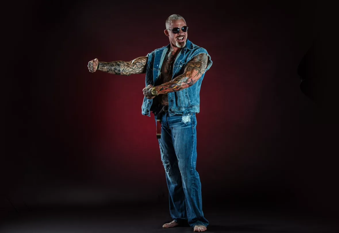 Rick Koenig — dramatic studio portrait in denim vest and sunglasses, dark maroon-lit backdrop